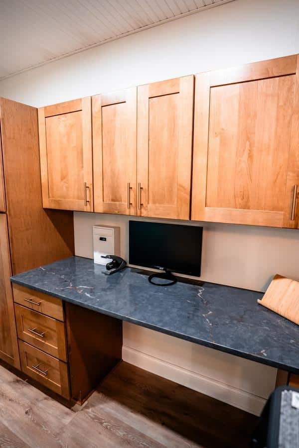 A home office space in the Spencerport Showroom features wooden cabinets, a black countertop, a computer monitor with headphones, a small safe, and a wooden cutting board on the right—all set against a light wall and wood floor.