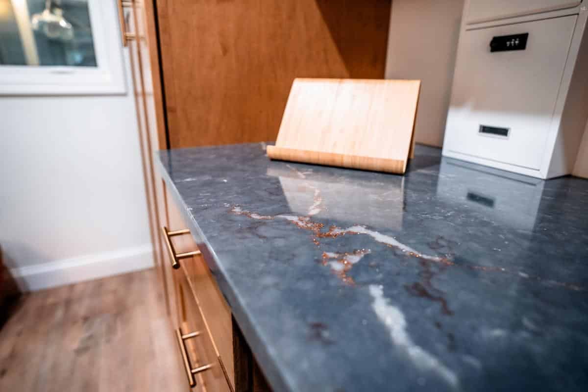 Close-up of a dark gray marble kitchen countertop with copper-colored veining, wooden cabinets with gold handles, and a white appliance in the background—discover elegant details like these at our Spencerport Showroom.