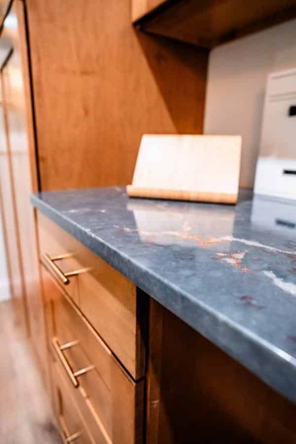 A close-up of a kitchen counter with a gray, marbled stone surface, wooden cabinets with gold handles, and a small wooden stand—captured in the elegant Spencerport Showroom.