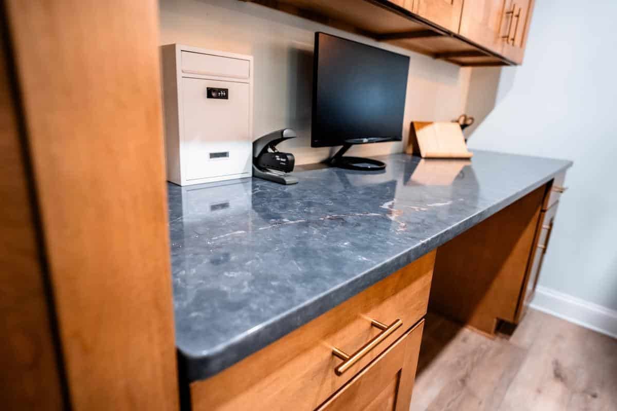 A home office desk with a gray stone countertop, wooden cabinets, a computer monitor, and sleek storage sits against a light-colored wall—discover similar modern setups at the Spencerport Showroom.