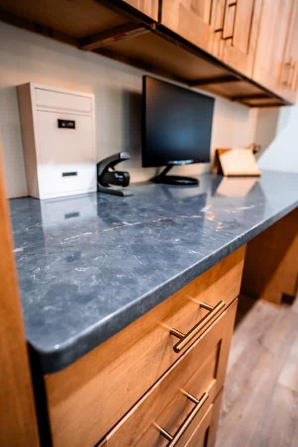 A modern office workspace in the Spencerport Showroom features wooden cabinets, a dark marble countertop, a drawer with gold handles, a computer monitor, a white file box, a headset, and a wooden stand on the counter.