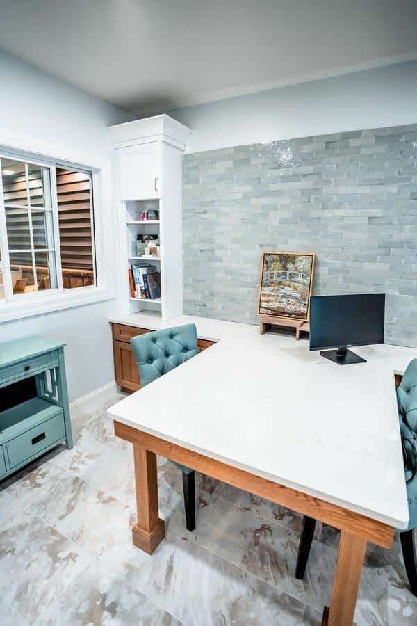Modern home office with a white marble desk, two teal tufted chairs, computer monitor, and bookshelf. An artwork is on display. Light tile floor and a pale green tiled accent wall add a fresh look inspired by the Spencerport Showroom.