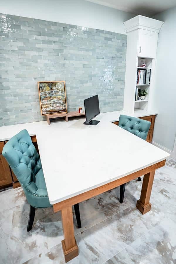 Modern office space with a white marble desk, two teal tufted chairs, a computer monitor, built-in shelves, and a light blue tiled accent wall. Artwork and books are displayed on the counter and shelves in the Spencerport Showroom.