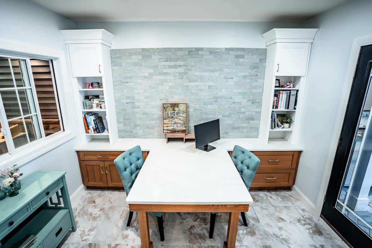 Modern home office with two teal upholstered chairs at a white marble desk, a computer monitor, wooden cabinets, shelves with books, a painting, and light blue tile backsplash. Large window and black door evoke the style of our Spencerport Showroom.
