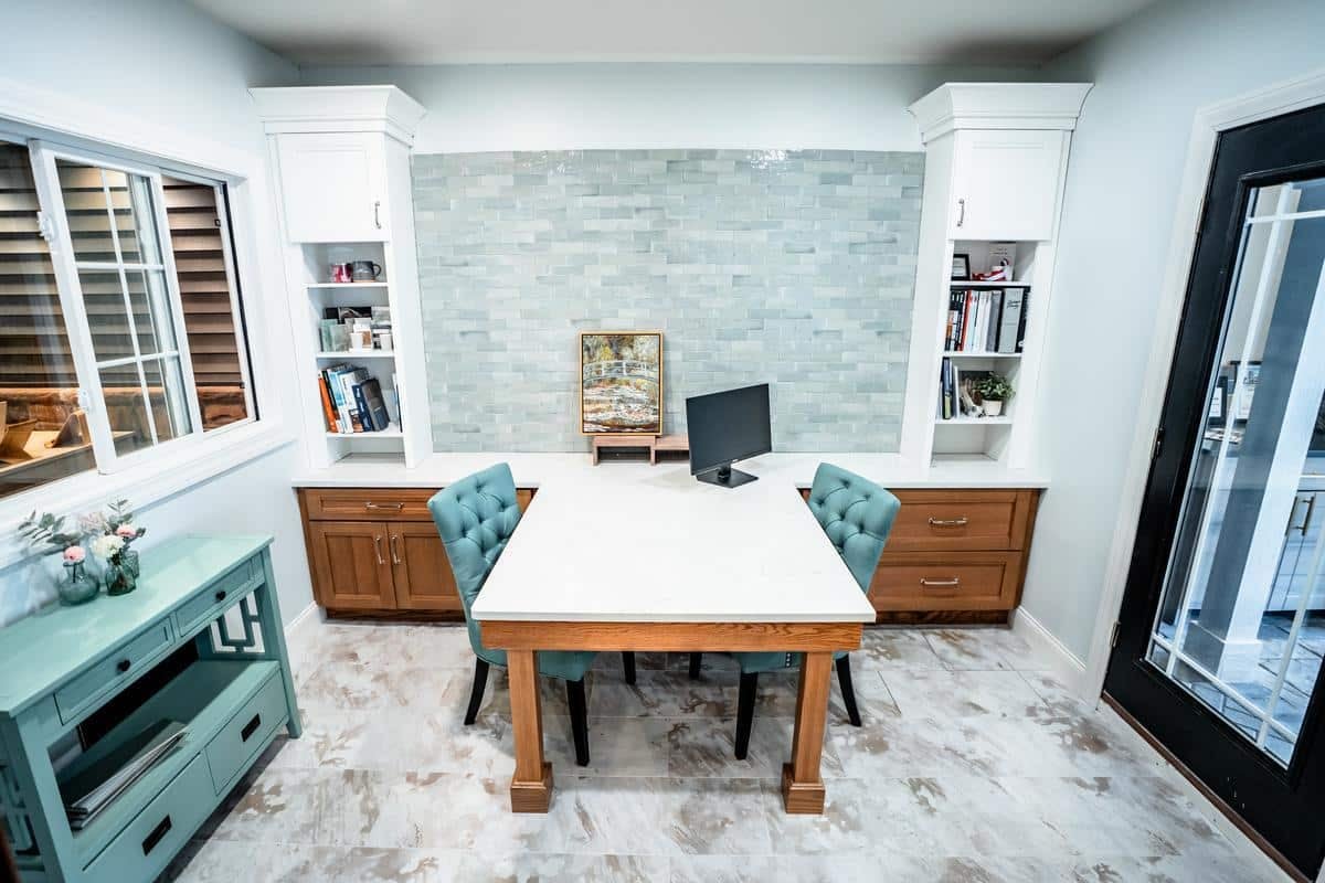 A modern home office with a marble-topped desk, teal chairs, wall shelving, a small monitor, decorative items, and a window—reflecting the elegant design seen in the Spencerport Showroom. The room has light-colored walls and tile flooring.