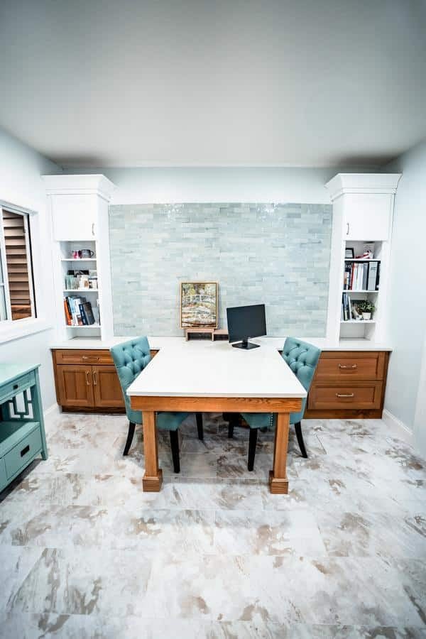 A modern home office with a wooden desk, two teal upholstered chairs, a computer monitor, built-in shelves with books and decor, and a light blue tile backsplash—showcasing inspiration from the Spencerport Showroom. White walls and a marble-patterned floor complete the space.