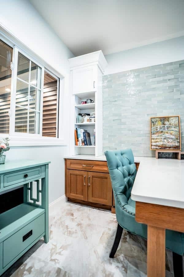 A cozy home office inspired by the Spencerport Showroom features a turquoise chair, wooden cabinets, a turquoise side table, a desk, and a tiled accent wall. Shelves display books and decor; a painting rests on the cabinet as sunlight streams in.
