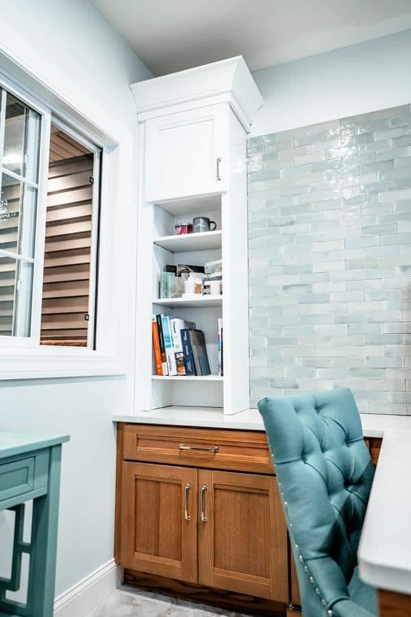 A built-in shelving unit with white upper shelves and wood lower cabinets, holding books, mugs, and other items, stands beside a window and a wall with glossy light blue tiles in the Spencerport Showroom. A teal tufted chair is in the foreground.
