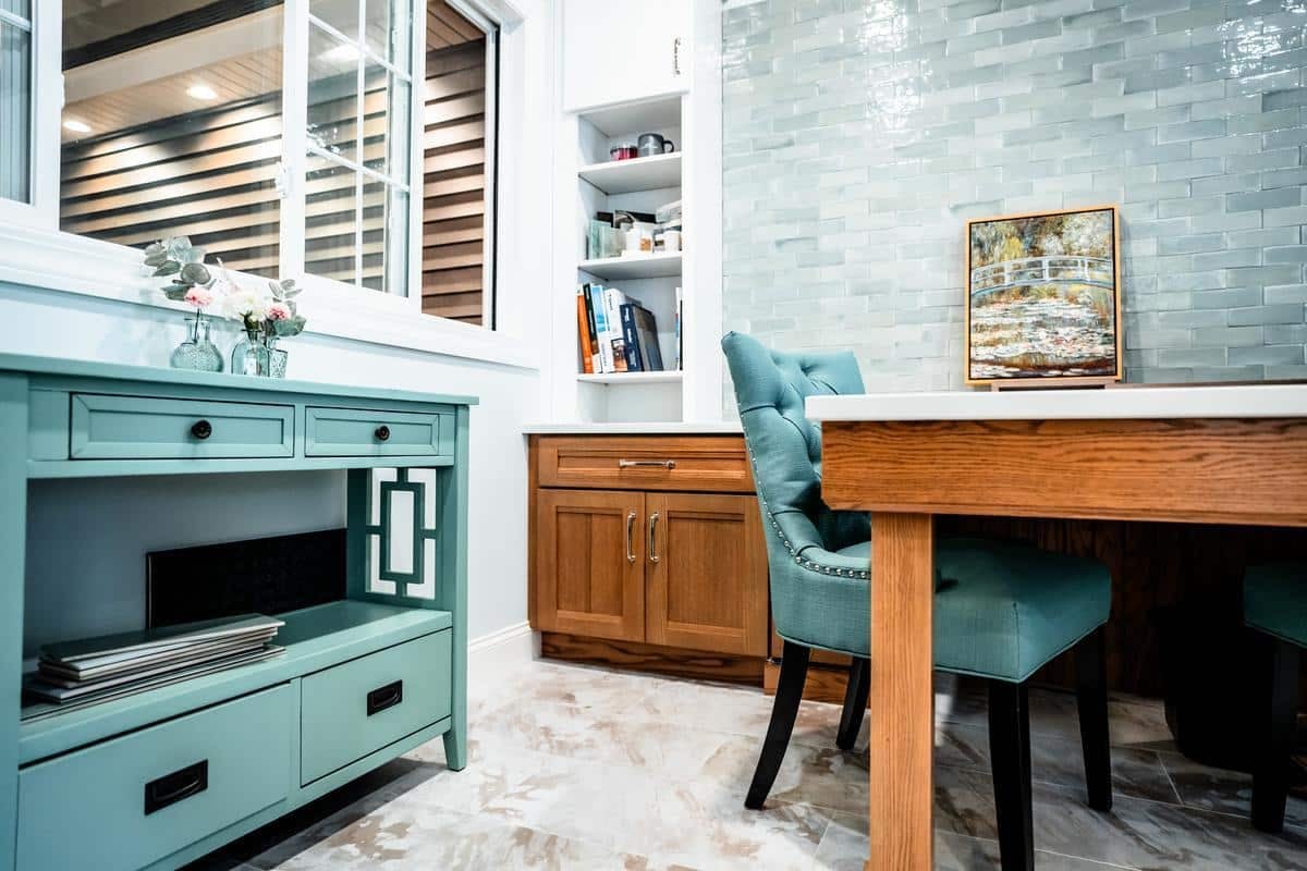 A cozy dining area in the Spencerport Showroom features teal chairs, a wooden table, a light blue cabinet, and book-filled shelves. Glossy light green tiles cover the wall, with a window looking out to another part of the house.
