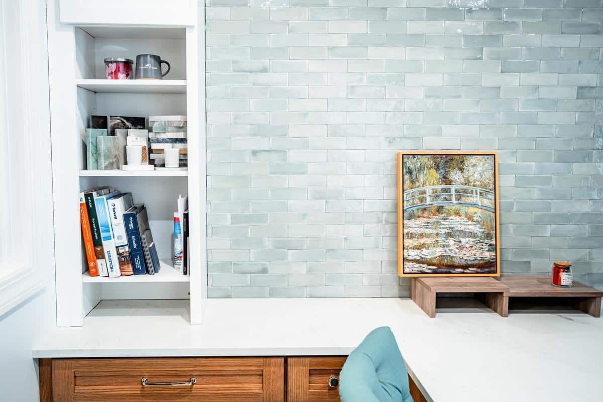 A modern kitchen corner in Spencerport features white shelves with books, mugs, and jars, a light gray subway tile backsplash, a framed painting of a bridge, wooden drawers, and a small candle on a wooden stand—like a stylish showroom display.