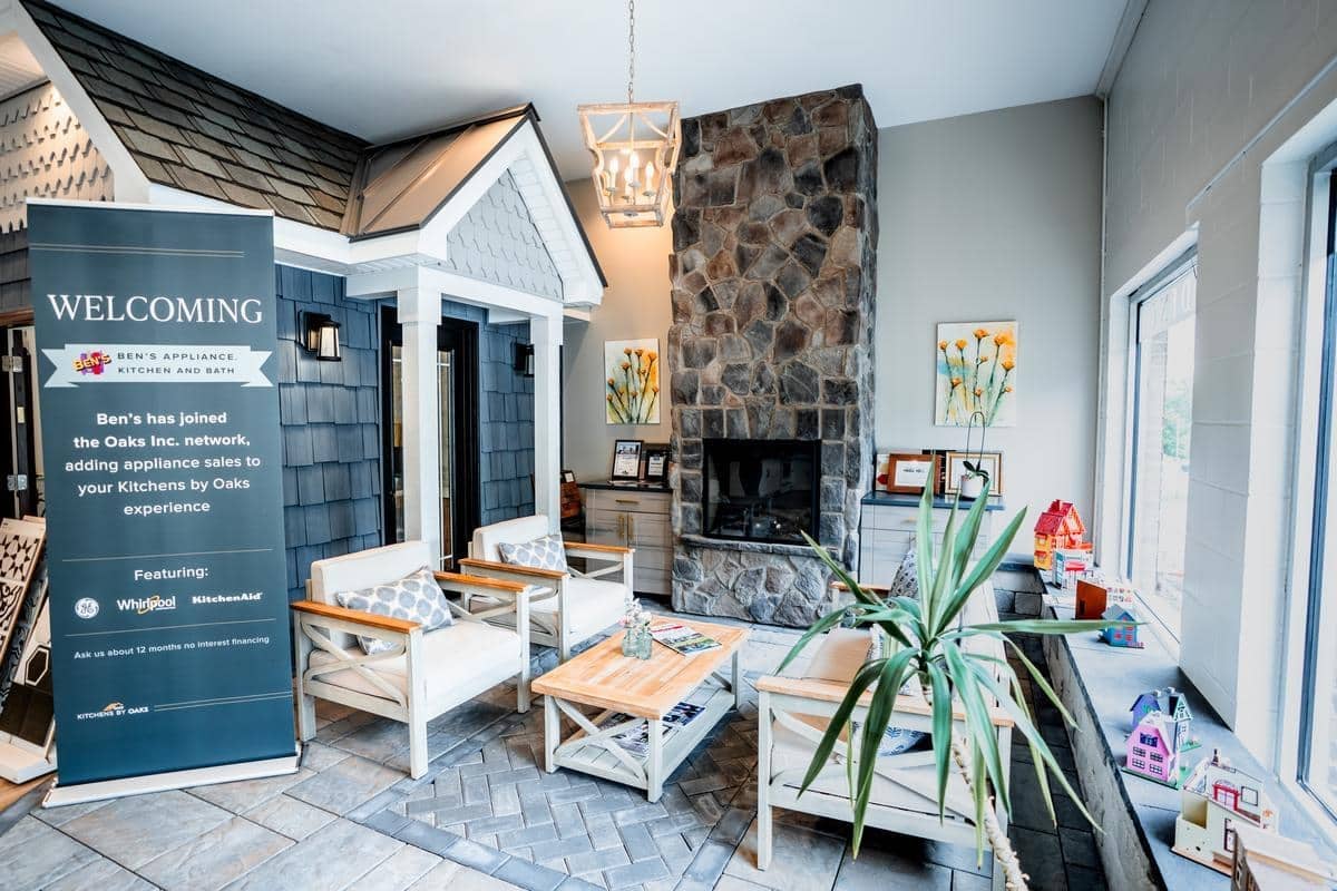 The Spencerport Showroom features a cozy stone fireplace, white and wood furniture, potted plants, floral artwork, and a welcome sign announcing a partnership with kitchen appliance brands. Large windows fill the space with natural light.