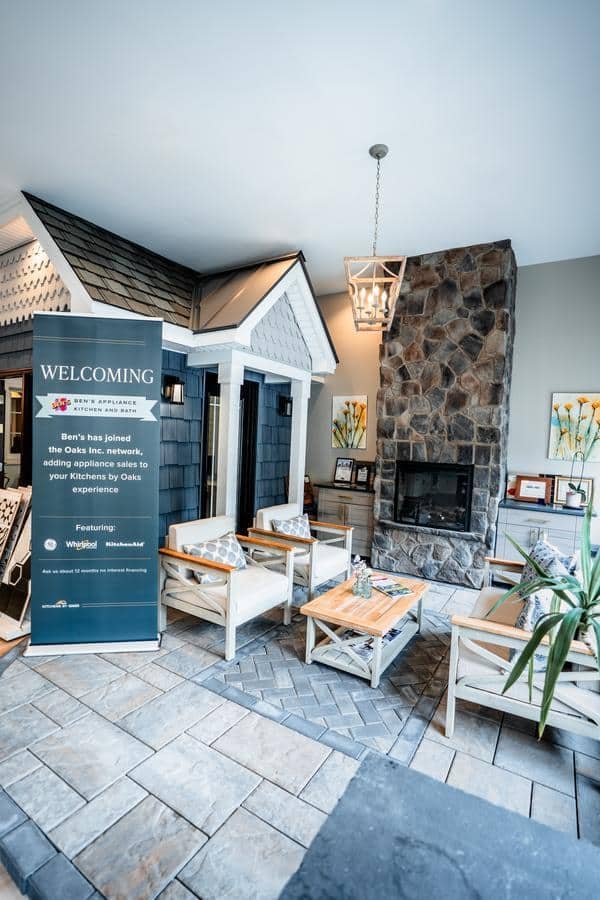 A cozy indoor seating area in this Spencerport showroom features white wooden chairs, a coffee table, a stone fireplace, and a pendant light. A welcoming banner and homey décor complete the space, bathed in natural light.