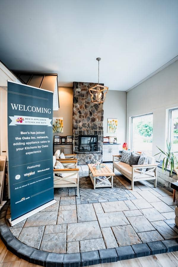 A cozy Spencerport Showroom waiting area features wooden chairs, a stone fireplace, modern lighting, and large windows. A blue welcome sign stands to the left, with natural light brightening the inviting space.