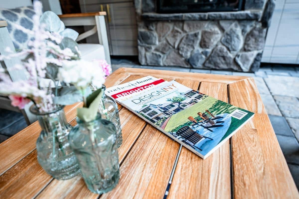 A magazine titled DesignNY rests on a wooden table next to two glass vases with flowers, in a cozy room with a stone fireplace—reminiscent of the inviting ambiance found in the Spencerport Showroom.