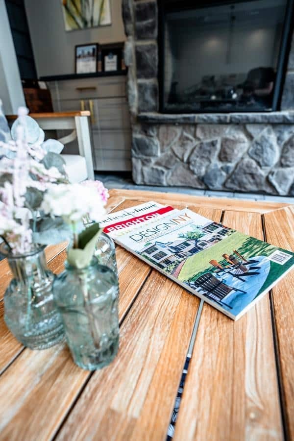A wooden table with glass vases holding white and pink flowers, next to a “Decor DIY” magazine, sits in a Spencerport showroom, with a stone fireplace and framed pictures in the background.