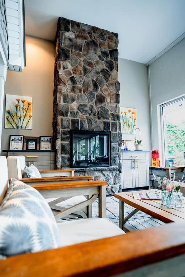 A cozy living room in Spencerport features a tall stone fireplace, wooden furniture with light cushions, floral artwork on the wall, and large windows. Magazines and flowers sit on a coffee table, evoking an inviting showroom feel.