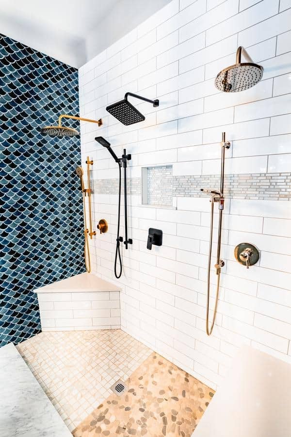 Modern shower with white subway tile walls, a section of blue fish scale tiles, multiple showerheads in black, gold, and silver finishes, and a floor with a mix of pebble and square tiles—featured in our Spencerport Showroom.