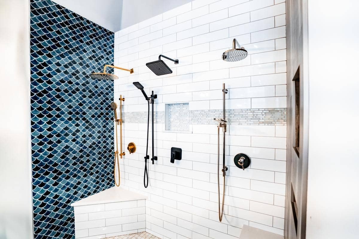 Modern shower with white subway tiles, a blue fish scale accent wall, multiple showerheads, and bronze and black fixtures. Built-in corner bench and niche shelf are also visible in our Spencerport Showroom.