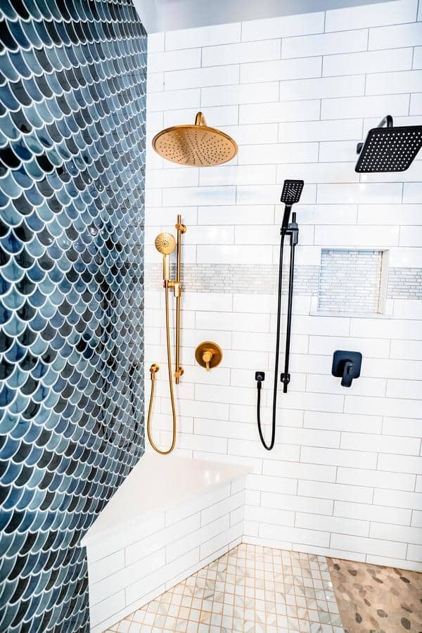Modern shower with white subway tiles, a blue fish scale accent wall, gold and black fixtures, built-in bench, and a niche with mosaic tile detail. Pebble tile flooring is also visible in this Spencerport Showroom display.