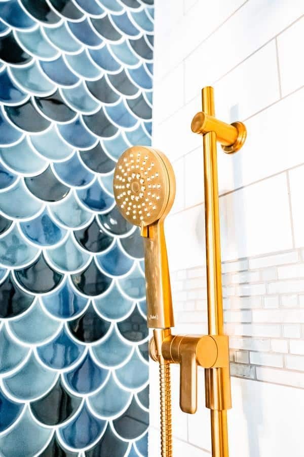 Close-up of a gold handheld shower head mounted on a rail, next to a wall with white subway tiles and blue fish scale pattern tiles, as seen in the Spencerport Showroom.