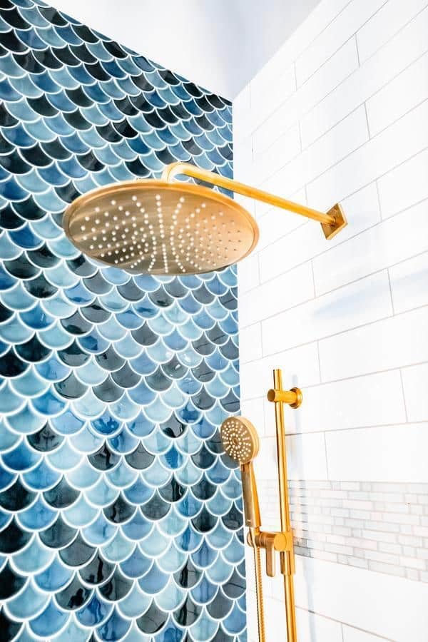 A modern shower with gold fixtures, including a large overhead rain shower and handheld showerhead, set against white subway tiles and blue fish scale pattern tiles—discover similar styles at the Spencerport Showroom.