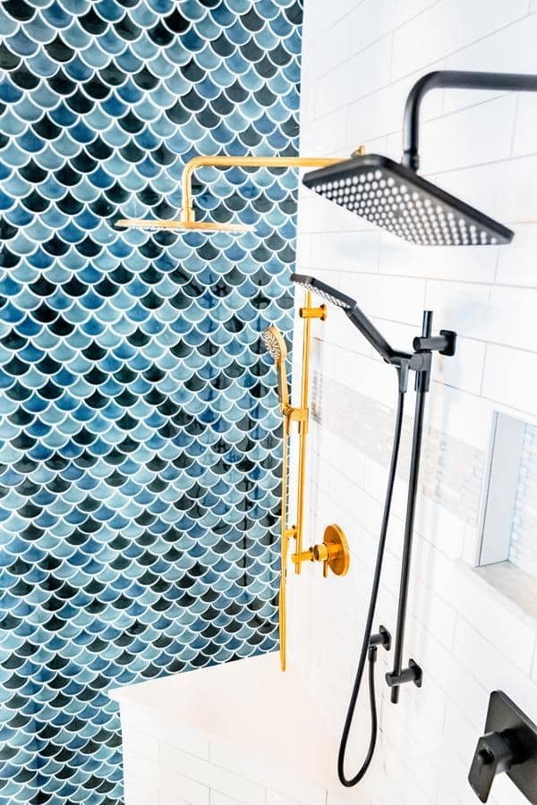 Modern shower with gold and black fixtures, set against a wall with blue fish scale and white rectangular tiles. A shower bench is visible in the corner—visit our Spencerport Showroom to experience this elegant design in person.