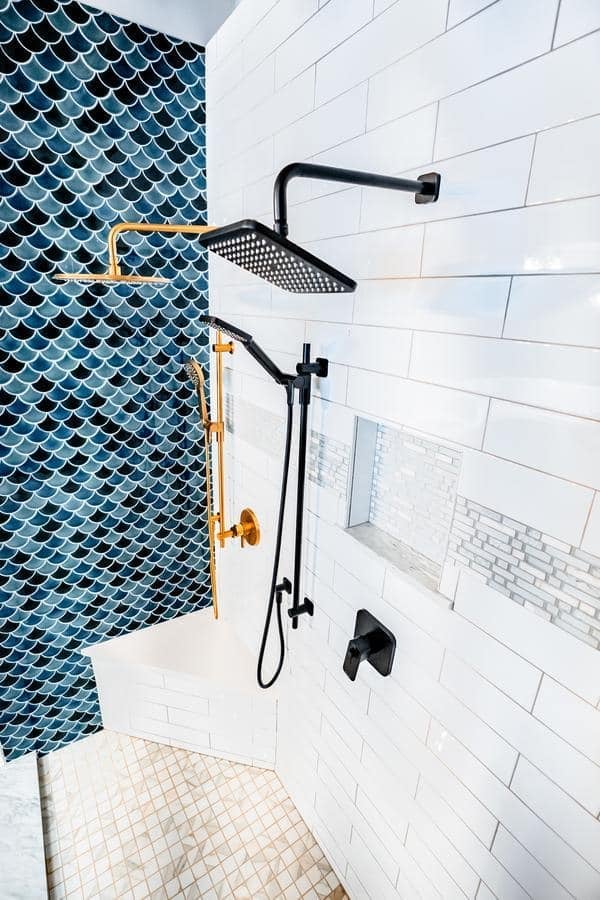 Experience a modern shower at the Spencerport Showroom, featuring white subway tiles, blue fish scale accents, a black square rainfall showerhead, gold and black fixtures, a built-in shelf, and a beige tiled floor.