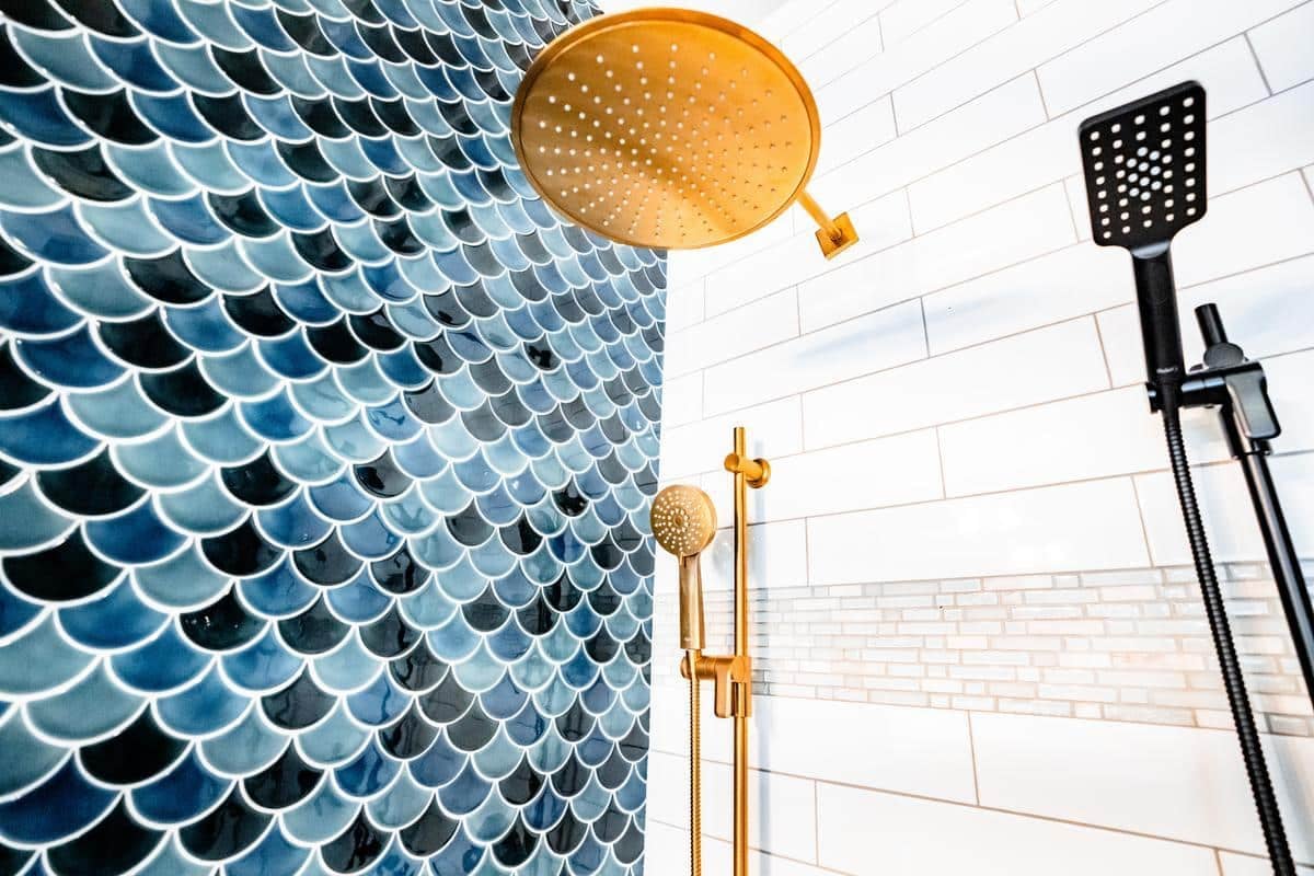 Close-up of a modern shower in a Spencerport showroom, featuring a gold rain showerhead, black handheld shower, blue fish-scale tiles on one wall, and white subway tiles with a mosaic accent on the other.