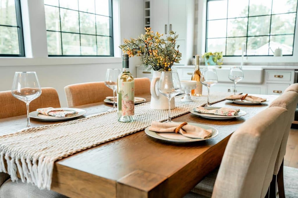 A dining table set for four with beige chairs, wine glasses, plates with napkins, a bottle of white wine, and a white vase with yellow flowers in a bright, modern Webster Showroom kitchen.