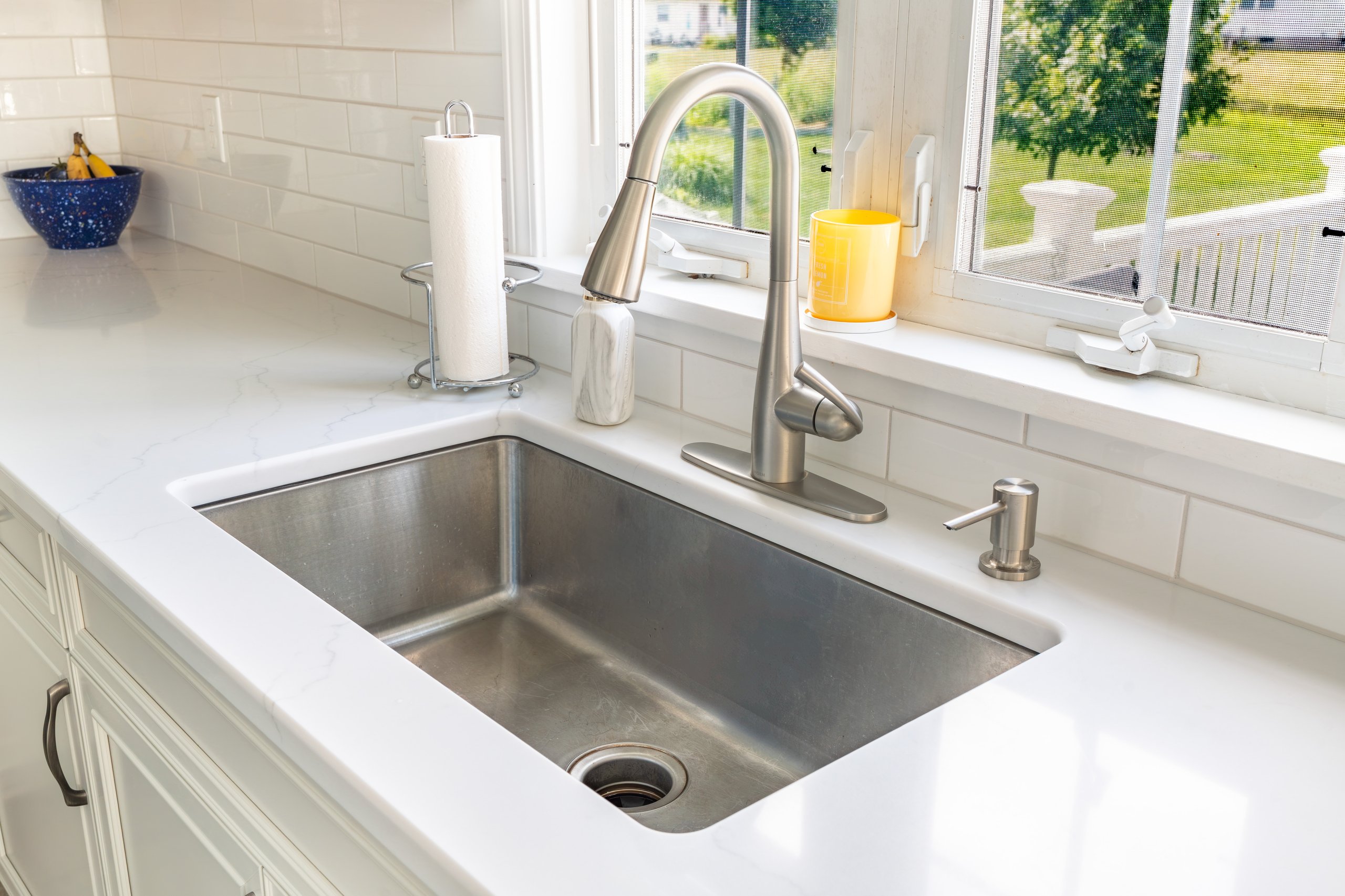 A clean stainless steel kitchen sink with a modern faucet, reverse osmosis system, soap dispenser, and paper towel holder on a white countertop. A yellow cup and sponge rest on the windowsill above, sunlight streaming in.