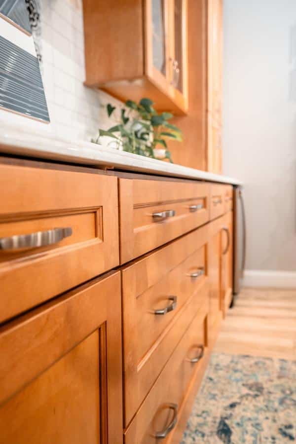 Close-up view of wooden kitchen cabinets with silver handles, a white countertop, and a small plant. The background features a backsplash and a patterned rug on light wood flooring, showcasing the style found in our Spencerport Showroom.