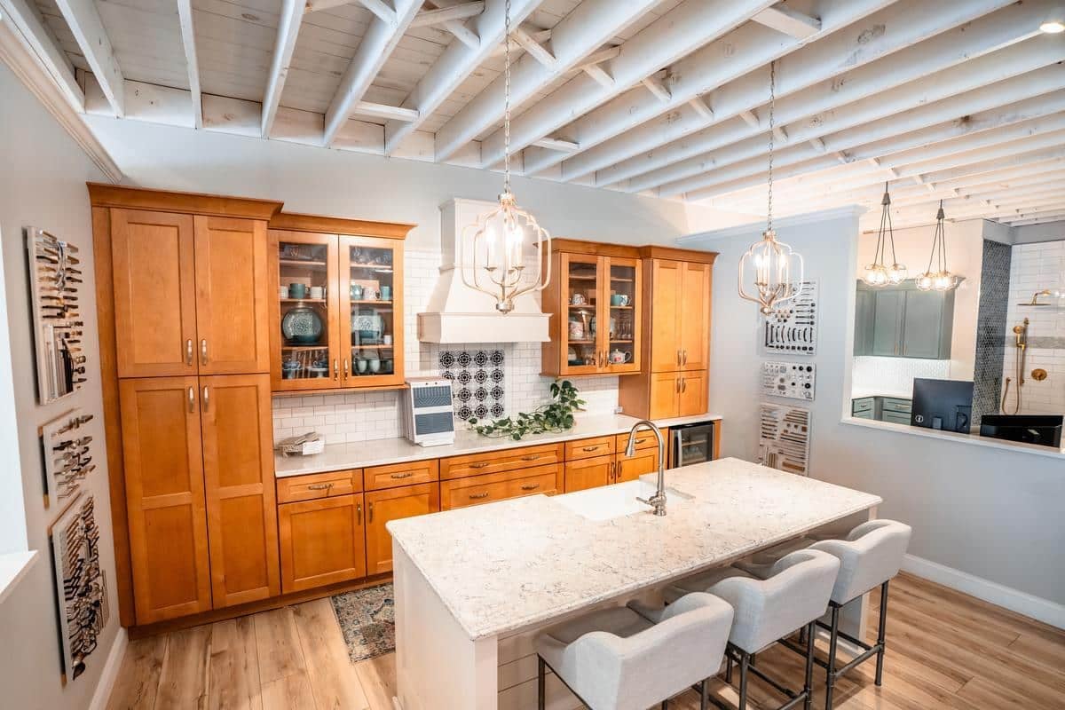 Modern kitchen with light wood cabinets, a white marble island with four cushioned stools, pendant lights, exposed ceiling beams, stainless steel appliances, and decorative wall art—featured at the Spencerport Showroom.
