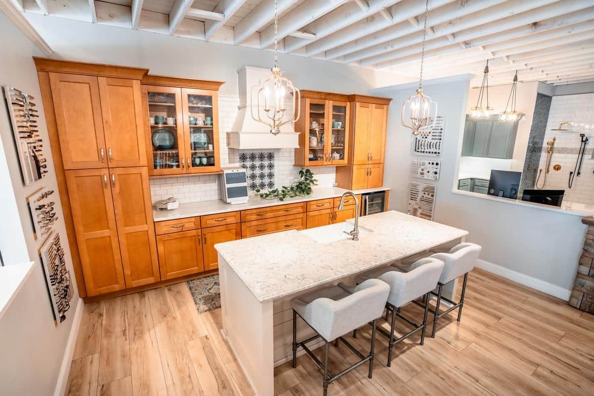 Modern kitchen with wooden cabinets, a marble island with three upholstered stools, pendant lights, and open ceiling beams—just like those featured in our Spencerport Showroom. Glass-front cabinets display dishes, and greenery decorates the countertop.