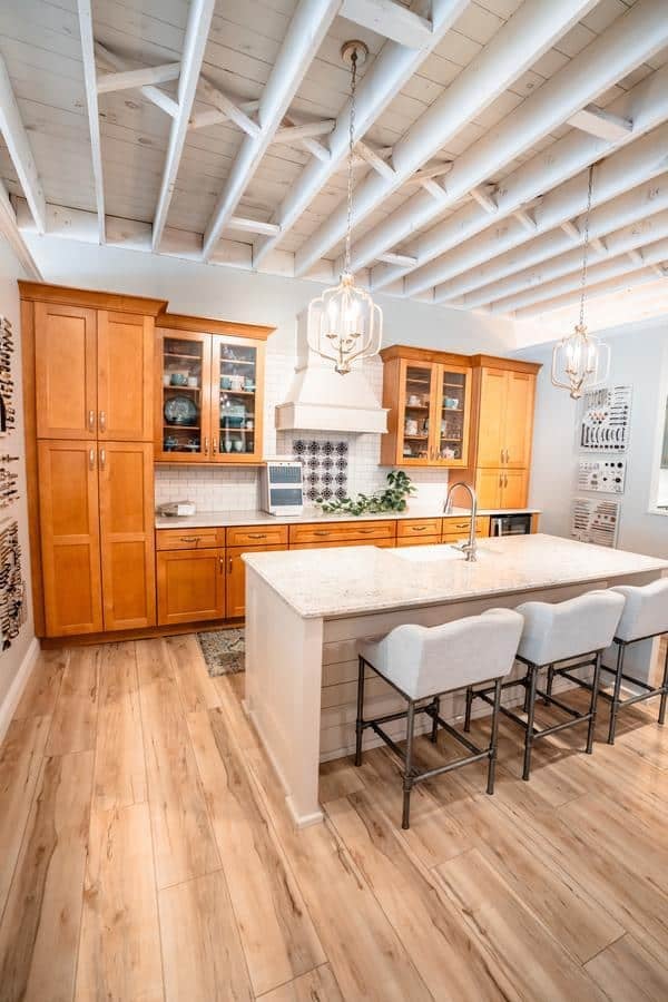 Modern kitchen with light wood cabinets, a large marble island with three upholstered stools, pendant lighting, exposed white ceiling beams, and light wood flooring. Visit our Spencerport Showroom to see this bright and neatly organized space in person.