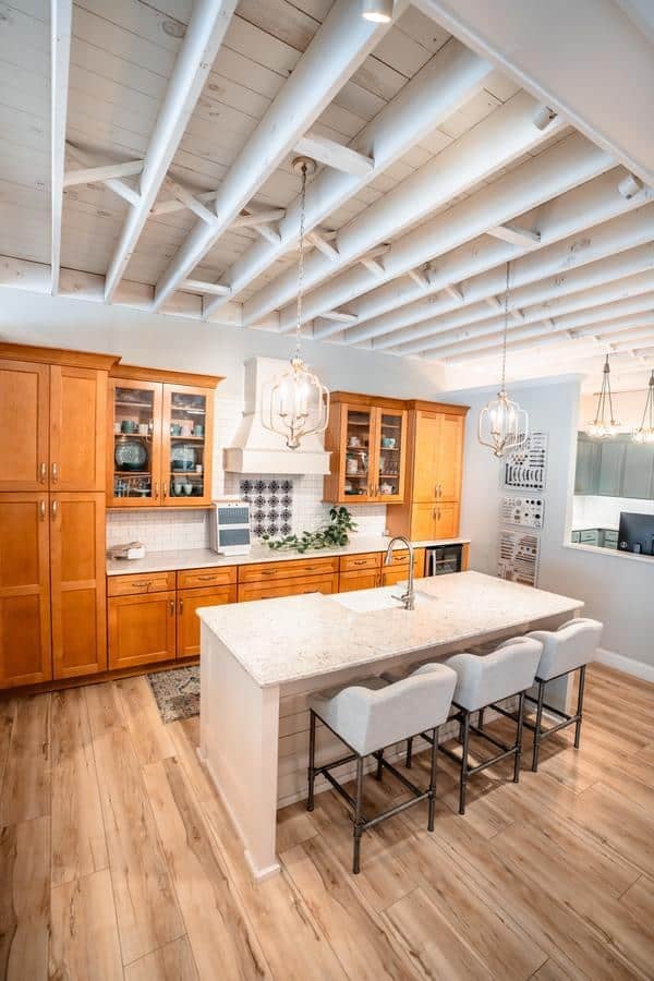 Modern kitchen with wooden cabinets, a marble island, three upholstered bar stools, pendant lights, and exposed white ceiling beams. Light wood flooring adds to the bright, airy feel—discover inspiration like this at the Spencerport Showroom.