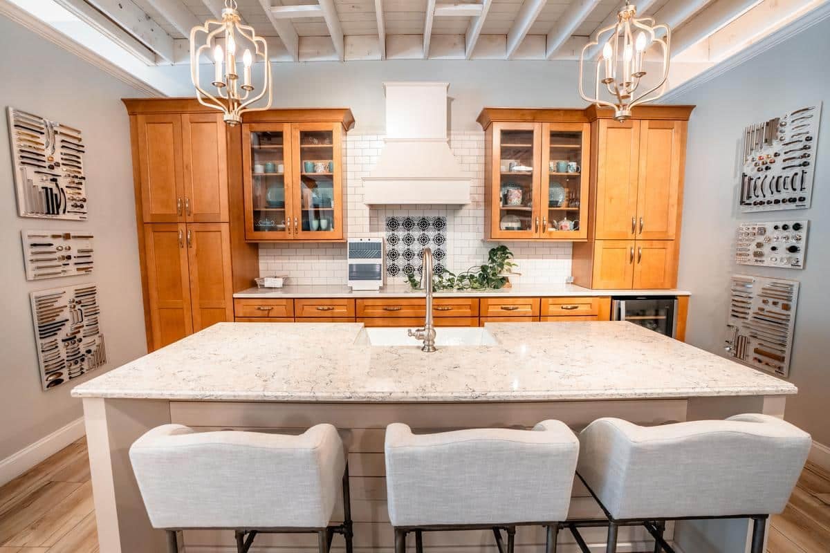 Modern kitchen in the Spencerport Showroom featuring light wood cabinets, a white marble island with sink, three upholstered barstools, pendant lights, and decorative wall art. Upper cabinets have glass doors; backsplash is pristine white tile.
