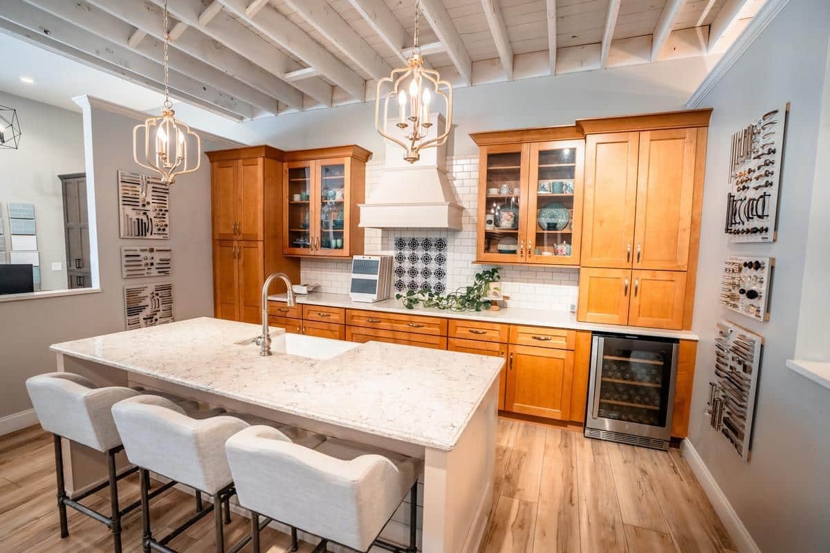 Modern kitchen with light wood cabinets, white marble island with sink, three upholstered stools, pendant lights, exposed ceiling beams, wine fridge, and decorative wall art—perfect inspiration from our Spencerport Showroom.