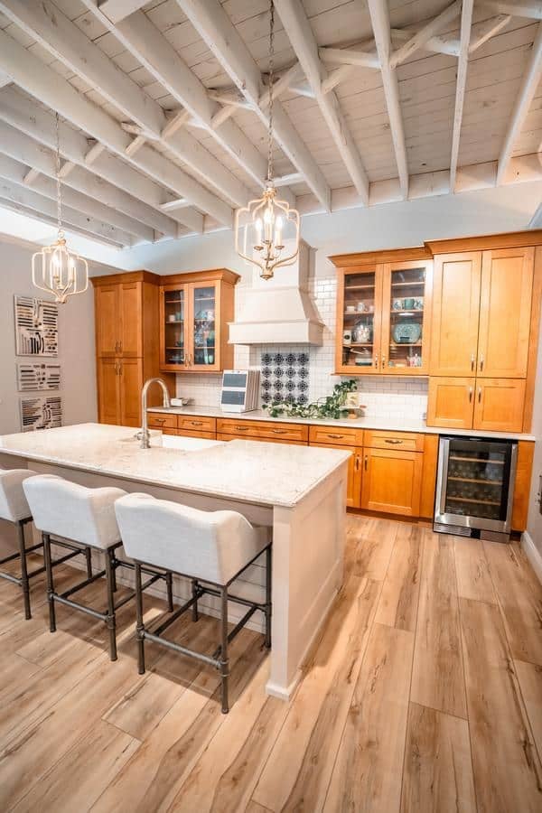 A modern kitchen in the Spencerport Showroom features light wood cabinets, a white marble island with three upholstered bar stools, pendant lights, exposed white ceiling beams, and light wood flooring. Greenery decorates the countertop.