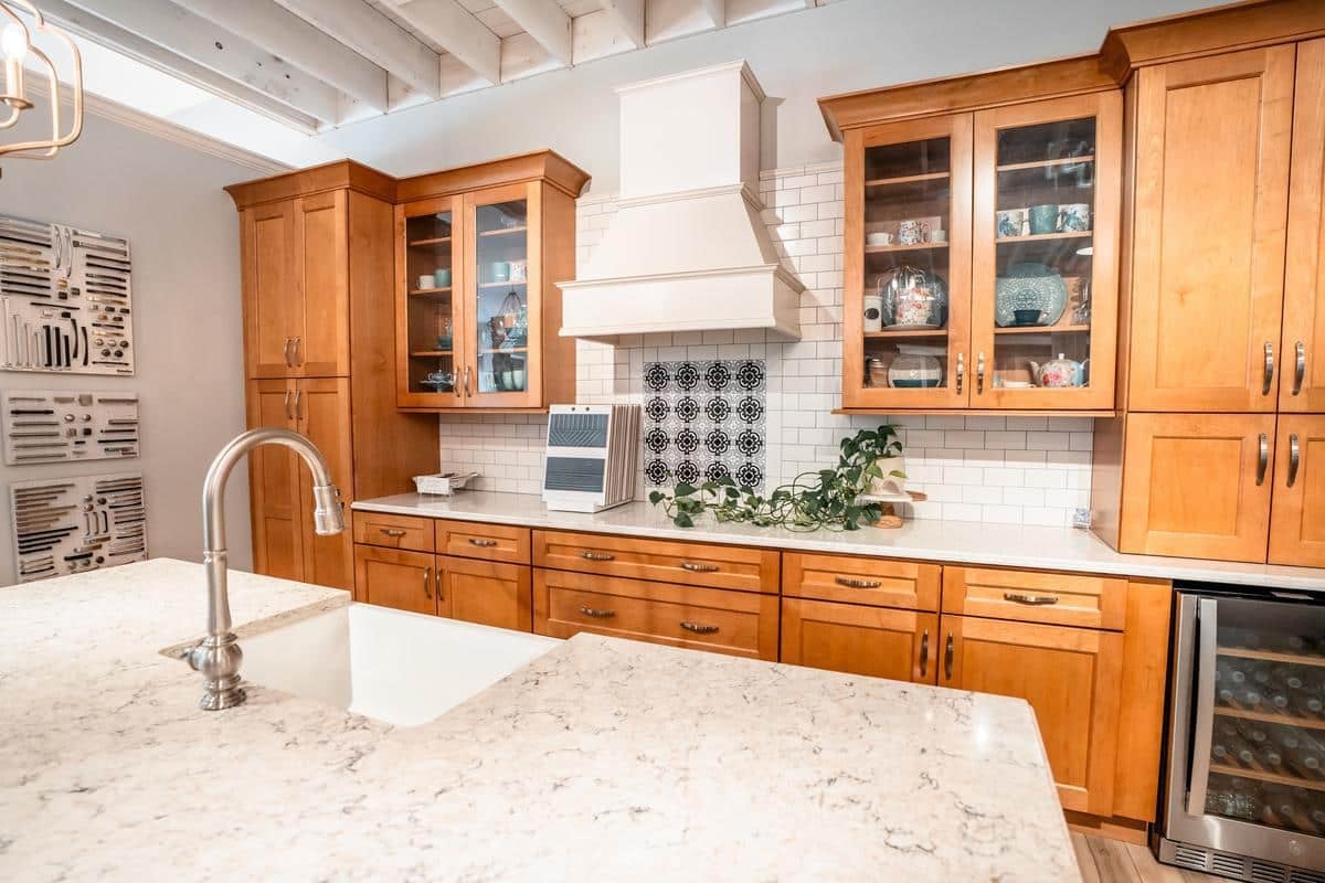 A modern kitchen with light wood cabinets, white marble countertops, a farmhouse sink, and glass cabinet doors is featured in the Spencerport Showroom. A wine fridge is built into the lower cabinets beneath a decorative tile backsplash.