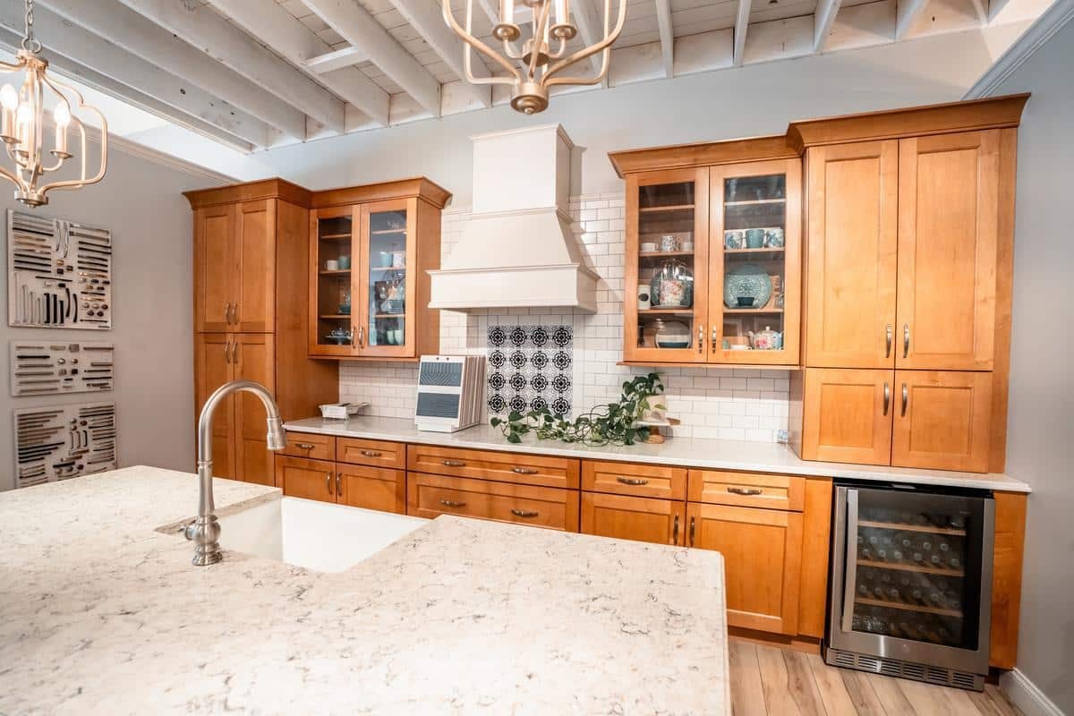 A modern kitchen with light wood cabinets, a white tile backsplash, marble countertops, and a wine fridge—just like those featured in our Spencerport Showroom. The island boasts a farmhouse sink and gooseneck faucet under decorative lighting.