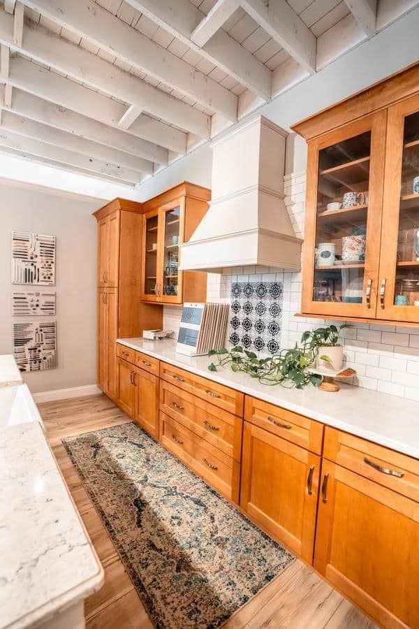 A bright kitchen with wooden cabinets, white countertops, a patterned backsplash behind the stove, glass-front uppers, a potted plant, and a runner rug on light wood floors under an open-beam ceiling—perfect inspiration from the Spencerport Showroom.