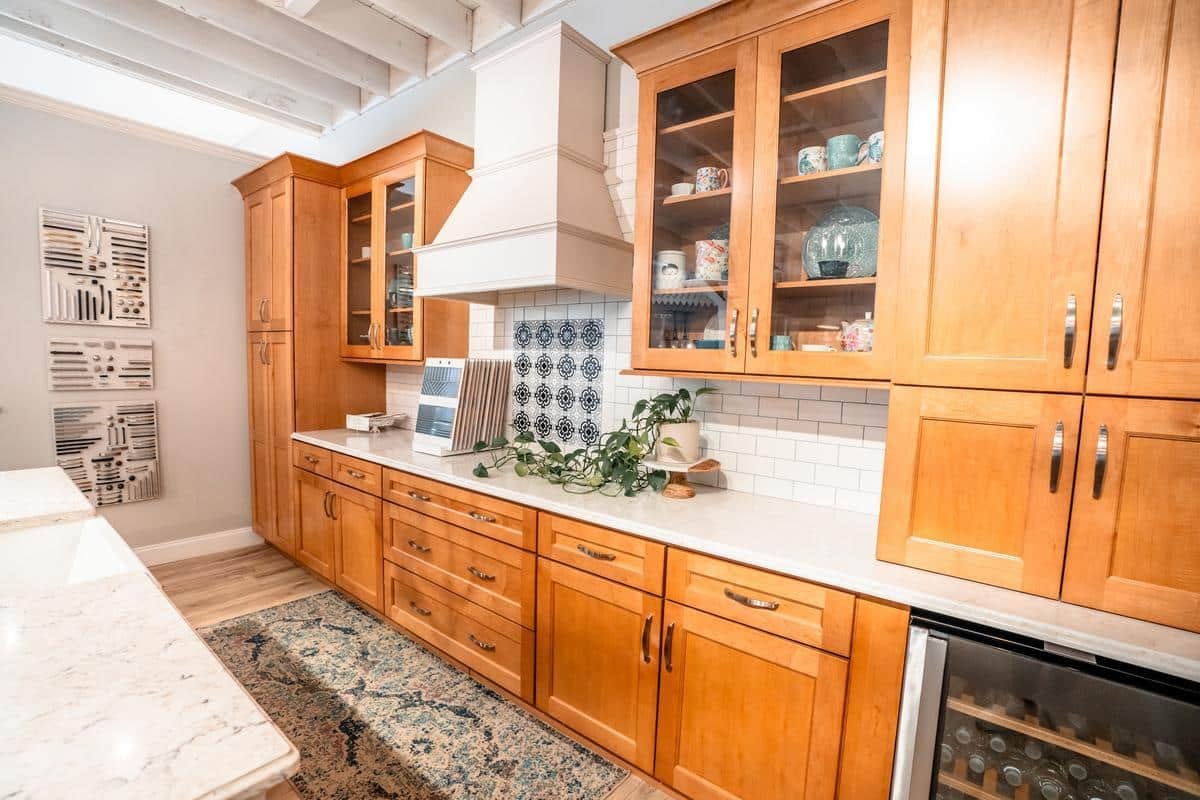 A modern kitchen with wooden cabinets, white countertops, a patterned rug, glass-front uppers, a white tile backsplash, and a small plant on the counter. Decorative wall pieces and a wine fridge complete this inviting Spencerport Showroom look.