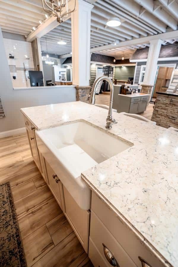 Modern Spencerport Showroom featuring a large farmhouse sink set in a white marble countertop, light wood flooring, and open ceiling beams. Various cabinetry and appliances are visible in the background.