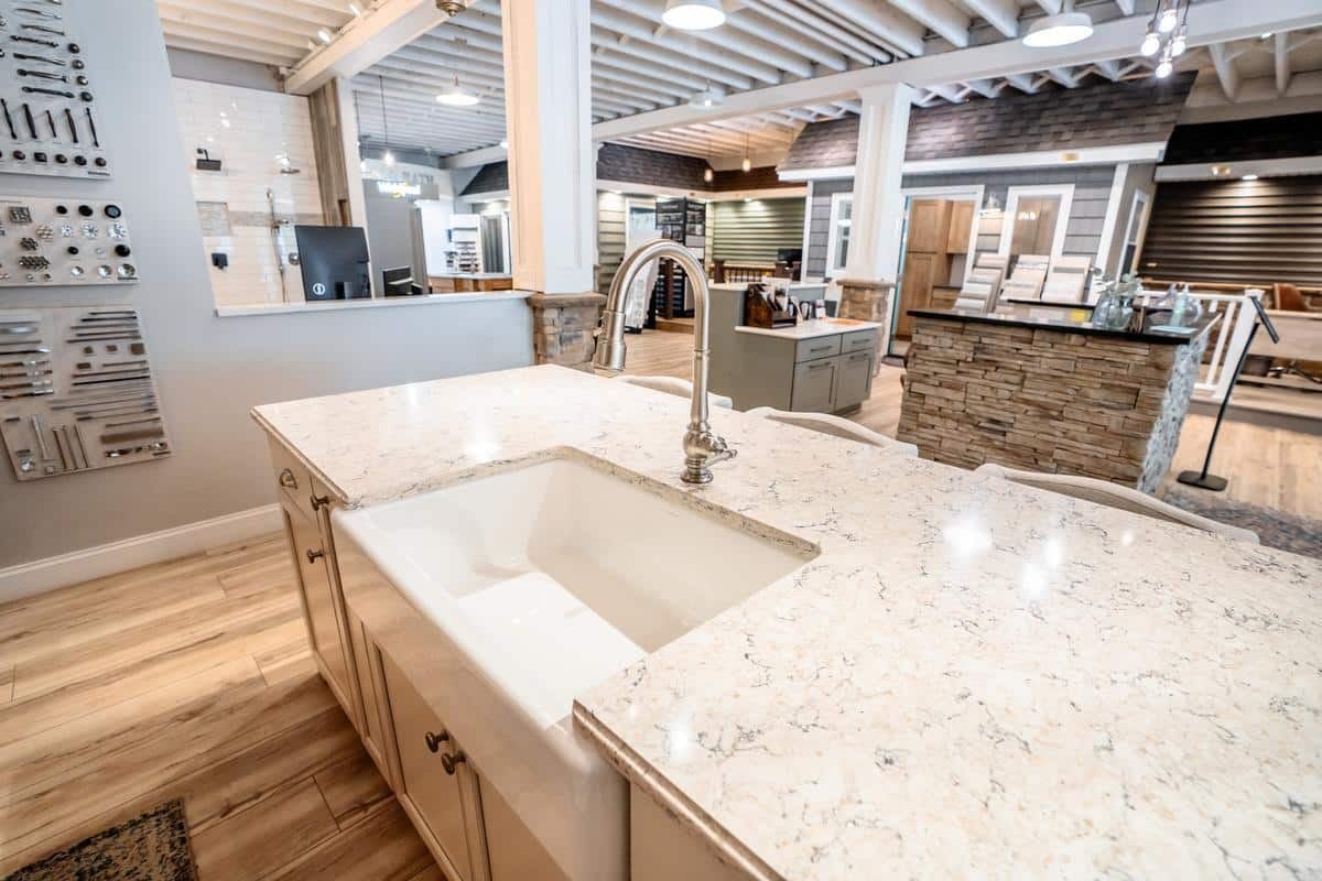 Marble countertop kitchen island with a built-in white farmhouse sink and silver faucet in the bright, modern Spencerport Showroom displaying cabinets, hardware, and home fixtures.