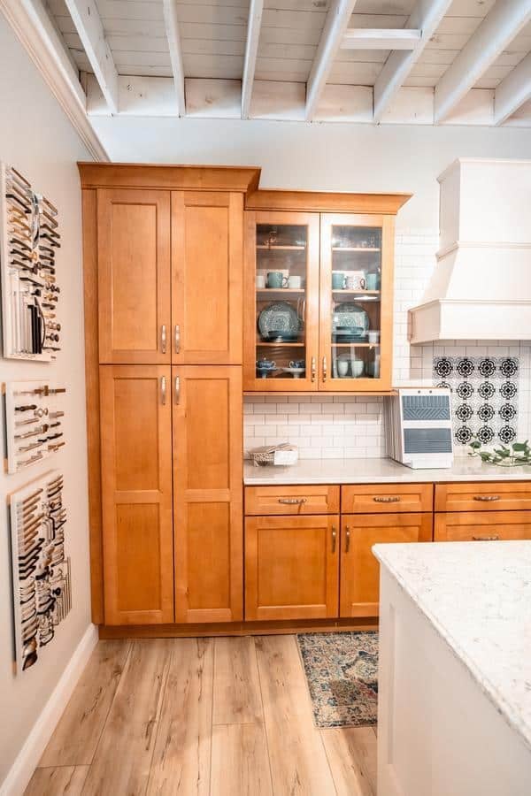 A kitchen with light wood cabinets, white subway tile backsplash, glass-front upper cabinets displaying dishes, and a countertop with a drying rack is showcased in the Spencerport Showroom alongside colorful patterned wall art featuring utensils.