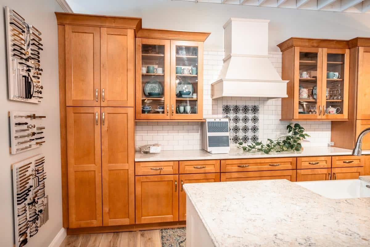 A kitchen with light wood cabinets, glass doors displaying dishes, a white range hood, white subway tile backsplash, black-and-white tile accent, knife racks on the wall, and a marble island countertop—just like our Spencerport Showroom.