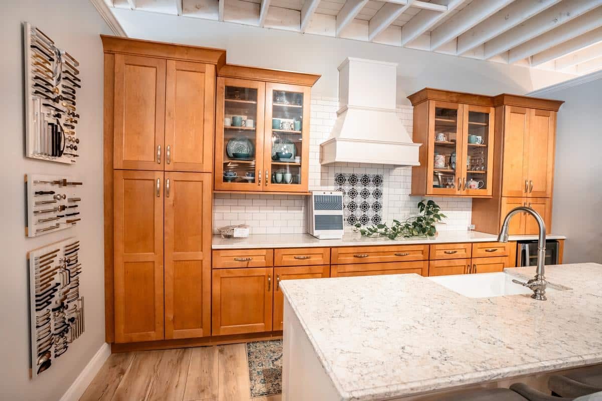 A modern kitchen features light wood cabinets, a white marble island with a sink, open shelving displaying dishes, and a wall-mounted knife rack—styles you can explore at the Spencerport Showroom. Decor includes a potted plant and patterned tile accents.