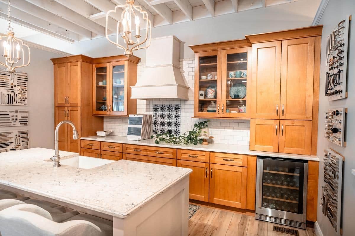 A modern kitchen with wooden cabinets, glass cabinet doors, a white tile backsplash, a marble island with a sink, and decorative lighting fixtures can be found at the Spencerport Showroom. Includes built-in wine cooler for added style.