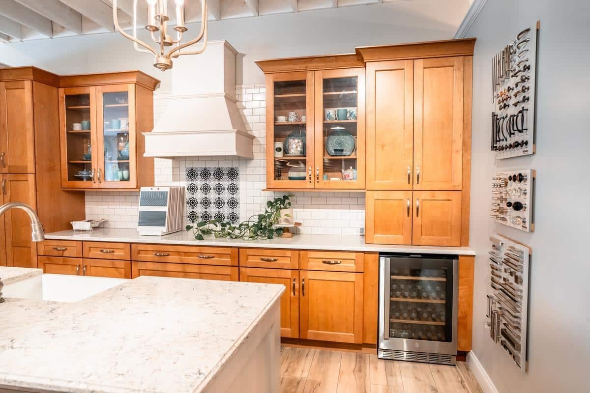 A bright Spencerport Showroom kitchen features wooden cabinets, a white island with a sink, glass-front cupboards, white subway tile backsplash, wine cooler, potted plant, and decorative wall art. Dishes and bowls are visible in the cabinets.