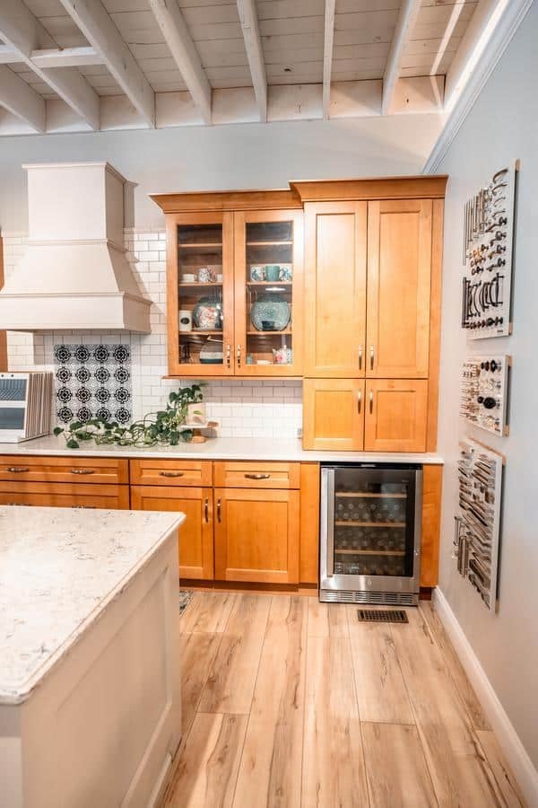 A modern kitchen with light wood cabinets, a glass-front display cabinet, wine fridge, white subway tile backsplash, and wall-mounted utensil racks. Featured in our Spencerport Showroom, the island has a striking white countertop in the foreground.